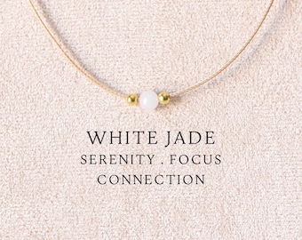 May include: A delicate necklace featuring a thin beige cord, a single white jade bead, and two small gold-toned metal beads. The text WHITE JADE SERENITY . FOCUS CONNECTION is printed below the bead. The necklace is displayed on a beige fabric.