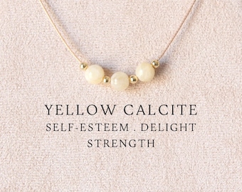 Puede incluir: Un collar delicado con tres cuentas redondas de color amarillo pálido separadas por pequeñas esferas metálicas doradas en un cordón fino. El texto YELLOW CALCITE SELF-ESTEEM . DELIGHT STRENGTH está impreso bajo el collar en la tarjeta.