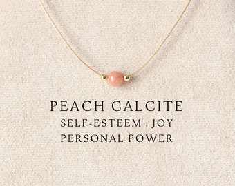 Puede incluir: Un collar delicado con una cuenta redonda de calcita melocotón centrada en un cordón fino. El texto debajo de la cuenta dice: PEACH CALCITE, SELF-ESTEEM . JOY, PERSONAL POWER. El collar se muestra sobre un soporte cubierto de tela beige.
