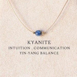 May include: A delicate necklace featuring a single round blue kyanite bead flanked by two small gold-toned metallic beads on a thin cord. The text KYANITE INTUITION . COMMUNICATION YIN-YANG BALANCE is printed below the necklace on the display.