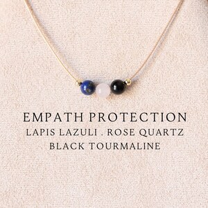 May include: A delicate necklace featuring three small beads: a blue lapis lazuli, a pale pink rose quartz, and a black tourmaline, strung on a thin cord. The text EMPATH PROTECTION, LAPIS LAZULI, ROSE QUARTZ, BLACK TOURMALINE is printed below.