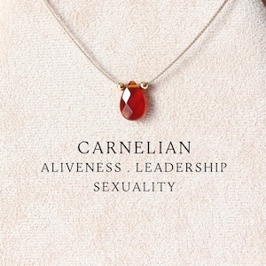 May include: A faceted, teardrop-shaped carnelian pendant on a thin, light-colored cord. The text below the pendant reads: CARNELIAN, ALIVENESS . LEADERSHIP, SEXUALITY. The stone is a deep, translucent orange-red color with small gold-toned beads.
