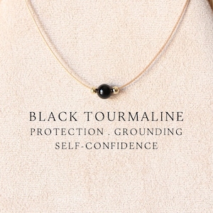 May include: A necklace featuring a single black tourmaline bead centred on a thin cord, flanked by two small gold-toned beads. The text BLACK TOURMALINE PROTECTION . GROUNDING SELF-CONFIDENCE is printed below the necklace on the display.