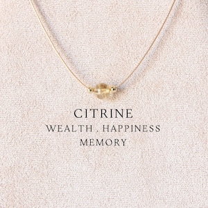 Könnte beinhalten: Eine zarte goldene Kettenkette mit einem einzelnen, hellgelben Citrin-Edelstein. Die Kette wird auf einem beigefarbenen, stoffbezogenen Ständer mit Holzsockel präsentiert. Der Text "CITRINE WEALTH. HAPPINESS MEMORY" ist unterhalb der Kette aufgedruckt.