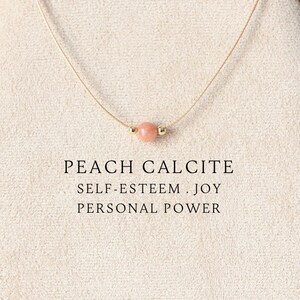 May include: A delicate necklace featuring a single round peach calcite bead centred on a thin cord. The text below the bead reads: PEACH CALCITE, SELF-ESTEEM . JOY, PERSONAL POWER. The necklace is displayed on a beige fabric-covered stand.
