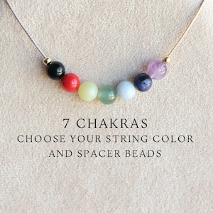 Puede incluir: Un collar con siete cuentas de chakra en los colores del arcoíris, con cuentas espaciadoras negras y doradas. El collar se muestra en un soporte de madera. 7 CHAKRAS ELIGE TU COLOR DE CUERDA Y CUENTAS ESPACIADORAS