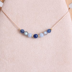 May include: A delicate necklace featuring a thin beige cord strung with seven round beads in shades of blue, gray, and iridescent stone, accented by small gold-toned metal spacers between the beads, creating a symmetrical arrangement.
