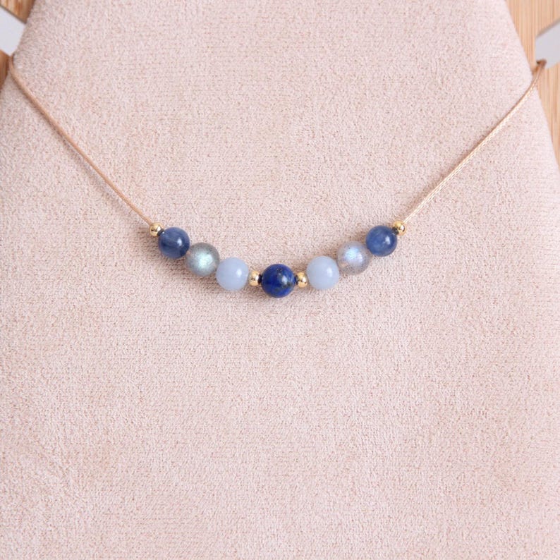 May include: A delicate necklace featuring a thin beige cord strung with seven round beads in shades of blue, gray, and iridescent stone, accented by small gold-toned metal spacers between the beads, creating a symmetrical arrangement.