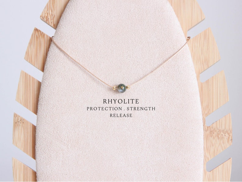 May include: A delicate necklace featuring a single, round rhyolite stone pendant. The stone is a mix of green and gray hues, suspended on a thin, beige cord. The text "RHYOLITE PROTECTION. STRENGTH. RELEASE" is displayed below the necklace.