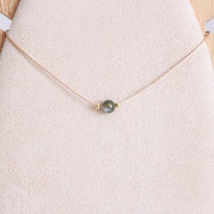 May include: A delicate necklace featuring a small, iridescent, dark green and gray bead as a focal point. The bead is accented with small gold-colored beads on either side. The necklace is strung on a thin, light-colored cord.