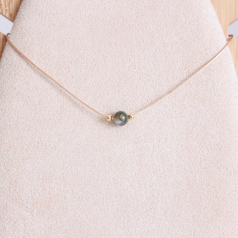 May include: A delicate necklace featuring a small, iridescent, dark green and gray bead as a focal point. The bead is accented with small gold-colored beads on either side. The necklace is strung on a thin, light-colored cord.