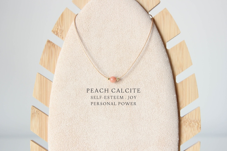 May include: A delicate necklace featuring a single round peach calcite bead centred on a thin cord. The text below the bead reads: PEACH CALCITE, SELF-ESTEEM . JOY, PERSONAL POWER. The necklace is displayed on a beige fabric-covered stand.