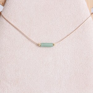 May include: A delicate necklace with a light green stone bar pendant and gold accents. The necklace is made with a thin, light brown cord.