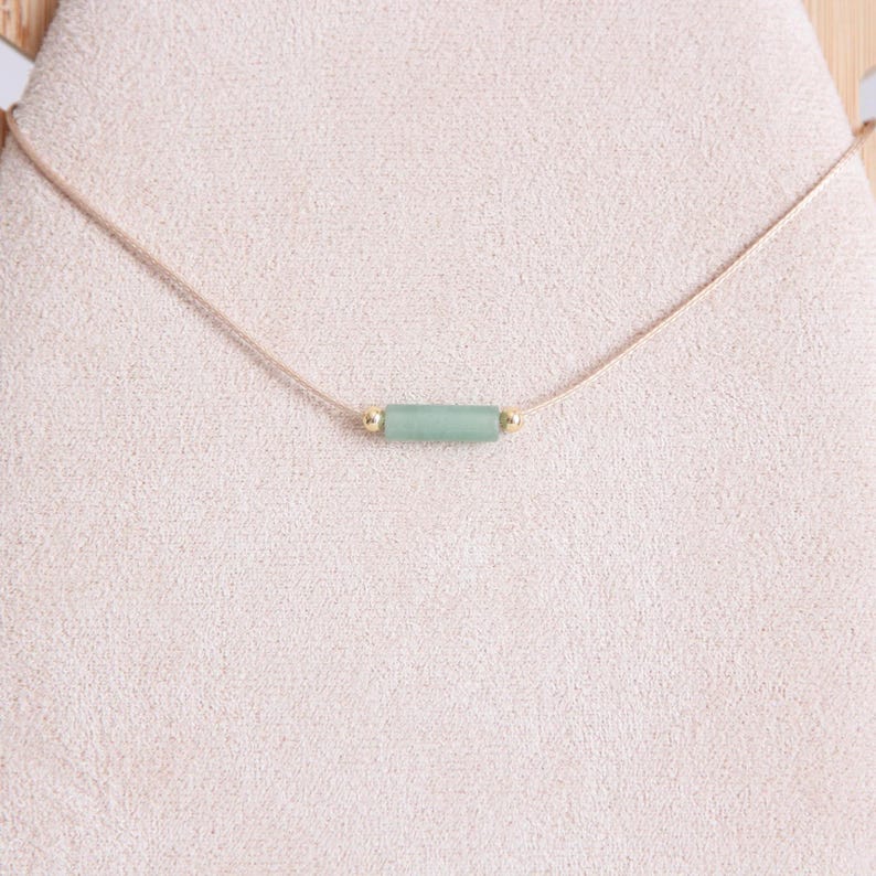 May include: A delicate necklace with a light green stone bar pendant and gold accents. The necklace is made with a thin, light brown cord.