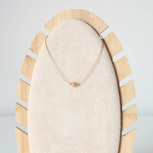 May include: A delicate necklace featuring a thin, light-coloured cord and a single, small, round, peach-coloured stone bead centred on the strand. The bead is flanked by two tiny, metallic gold-toned accents, creating a simple and understated design.