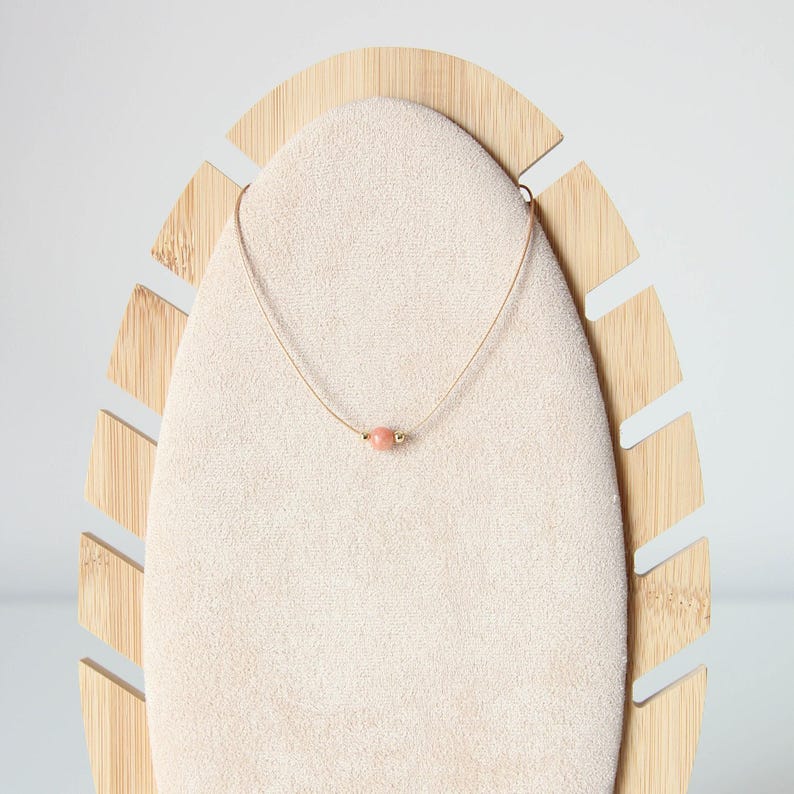 May include: A delicate necklace featuring a thin, light-coloured cord and a single, small, round, peach-coloured stone bead centred on the strand. The bead is flanked by two tiny, metallic gold-toned accents, creating a simple and understated design.