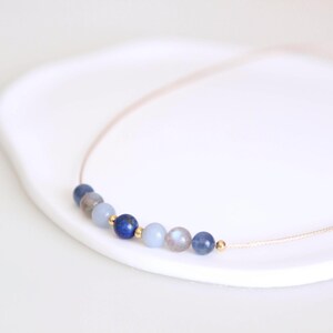 May include: A delicate necklace featuring a thin cord strung with a series of small, round beads in shades of deep blue, light blue, and grey, accented by tiny gold-toned metallic spheres interspersed between the stones along the center.