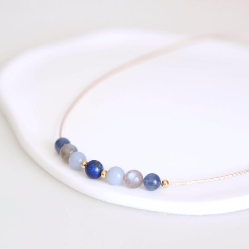May include: A delicate necklace featuring a thin cord strung with a series of small, round beads in shades of deep blue, light blue, and grey, accented by tiny gold-toned metallic spheres interspersed between the stones along the center.
