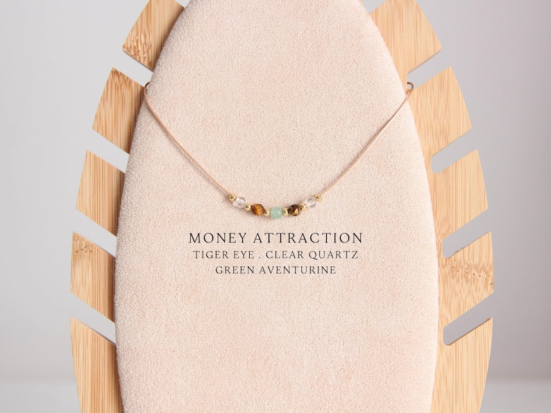 May include: A delicate necklace with a light brown cord and small beads of tiger eye, clear quartz, and green aventurine. The necklace is displayed on a wooden stand. The text "MONEY ATTRACTION TIGER EYE. CLEAR QUARTZ GREEN AVENTURINE" is written below the necklace.