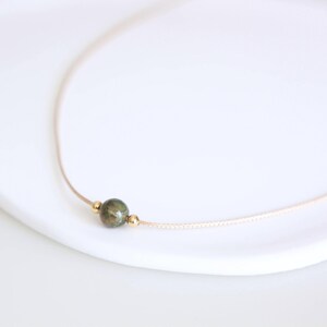 May include: A delicate necklace featuring a small, round, green stone pendant with gold-colored accents. The pendant is strung on a thin, beige cord. The necklace is displayed on a white surface.