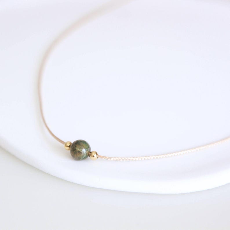 May include: A delicate necklace featuring a small, round, green stone pendant with gold-colored accents. The pendant is strung on a thin, beige cord. The necklace is displayed on a white surface.
