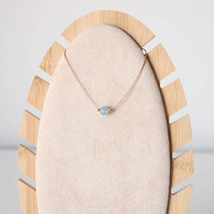 May include: A delicate necklace featuring a single, round, light blue gemstone bead centered on a thin, light-colored cord. The bead is flanked by two small, metallic gold-toned spheres, creating a simple and balanced design for this piece of jewelry.