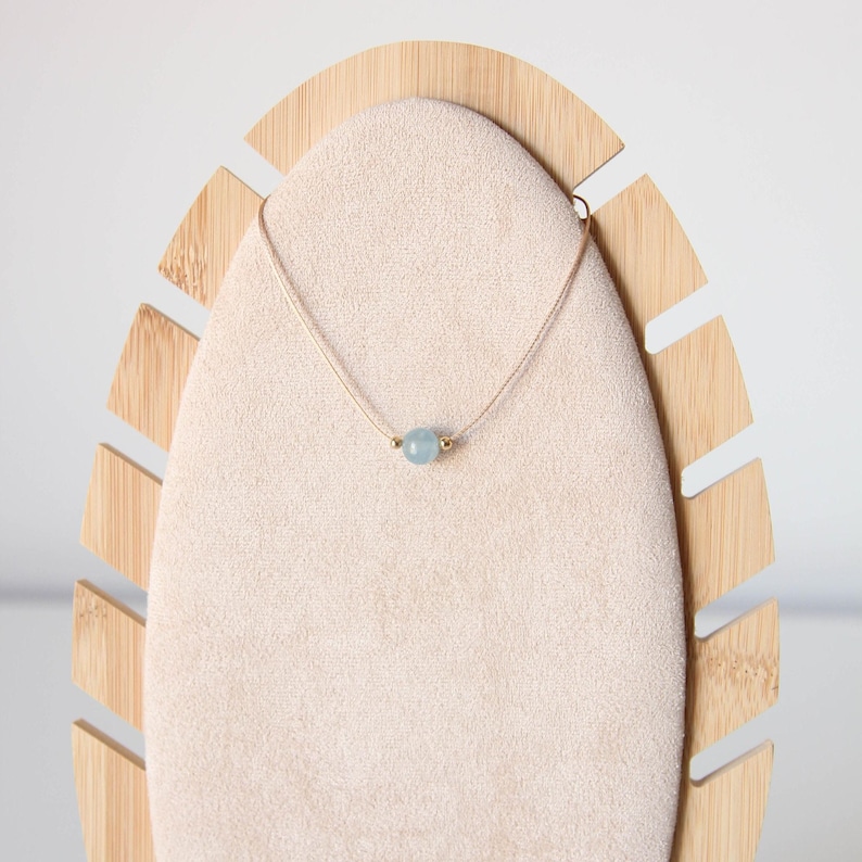 May include: A delicate necklace featuring a single, round, light blue gemstone bead centered on a thin, light-colored cord. The bead is flanked by two small, metallic gold-toned spheres, creating a simple and balanced design for this piece of jewelry.