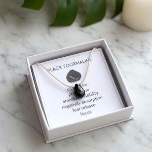 May include: A black tourmaline teardrop pendant on a light beige cord necklace, presented in a white gift box. The included card features the text BLACK TOURMALINE, a small stone graphic, and a list of properties including calm, protection, emotional stability, negativity absorption, fear release, and focus.