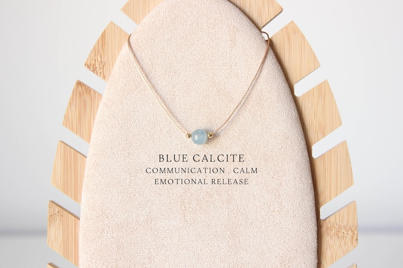 May include: A delicate necklace featuring a single light blue round gemstone bead flanked by two small gold-toned metal beads on a thin cord. The text BLUE CALCITE COMMUNICATION, CALM EMOTIONAL RELEASE is printed below the necklace on the display.