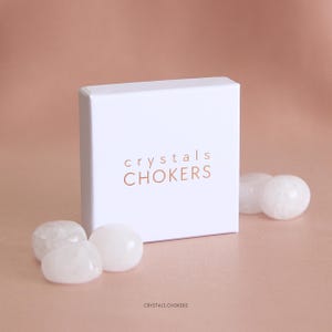 May include: A square white cardboard box with the text "crystals CHOKERS" printed in a copper-colored font on the front. The box is surrounded by several smooth, translucent, rounded white stones that appear to be polished quartz crystals.