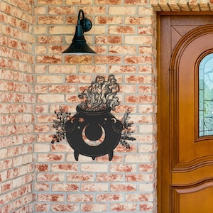May include: Black metal cauldron with a crescent moon and stars, surrounded by swirling smoke and floral accents, mounted on a brick wall next to a wooden door.