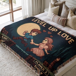 May include: A dark blue woven blanket with a pixelated video game design. The blanket features a couple holding hands under a full moon, with the text "LEVEL UP LOVE." The names "Daniel" and "Emily" are also displayed. The blanket has a fringed edge.