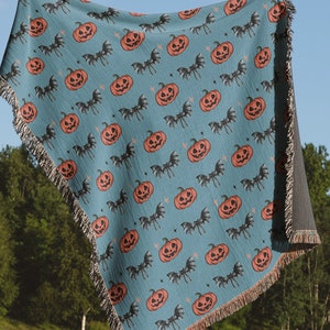 Op de afbeelding: Een geweven deken in tealblauw met een herhalend patroon van oranje pompoenen en zwarte vleermuizen. De deken heeft franjes aan alle kanten.