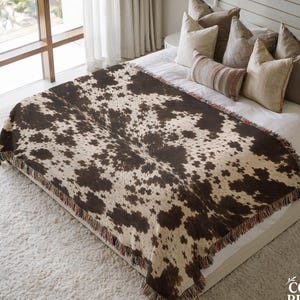 May include: A rectangular woven throw blanket featuring a brown and cream cowhide print pattern with fringed edges. The blanket is draped over a white bedspread, with the text THE COOL RULER CO. visible in the bottom right corner of the image.