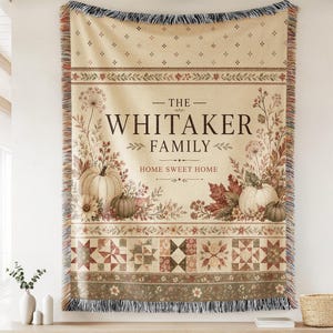 Könnte beinhalten: Eine gewebte Wohndecke mit cremefarbenem Hintergrund und herbstlichen Illustrationen von Kürbissen, Trockenblumen und Blättern. Der Text lautet THE WHITAKER FAMILY HOME SWEET HOME. Das Design umfasst eine Bordüre im Quilt-Stil und Fransen.