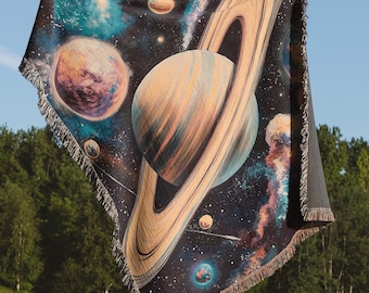 May include: A woven tapestry throw blanket featuring a colorful depiction of a galaxy with planets and stars. The blanket is held up in front of a person standing in a grassy field.