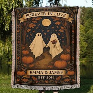 Op de afbeelding: Een geweven sprei met een romantisch Halloween-thema. Het ontwerp toont twee spookfiguren die elkaars hand vasthouden, pompoenen en de tekst "FOREVER IN LOVE" en "EMMA & JAMES EST. 2014". De deken heeft een zwarte rand en franjes.