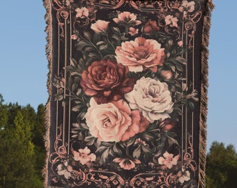 May include: A person holding up a large tapestry throw blanket with a floral pattern. The blanket is black with pink, red, and white roses and green leaves. The blanket has a fringed border.