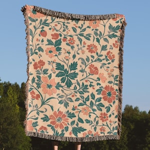 May include: A beige floral throw blanket with a repeating pattern of coral-colored flowers and teal leaves. The blanket has a fringed edge and is held up against a clear blue sky. The blanket is made of a woven material.