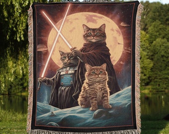 May include: A woven tapestry featuring three cats dressed as Star Wars characters. The cats are standing in front of a large moon with a starry background. The cat in the middle is dressed as Darth Vader, the cat on the left is dressed as a Jedi Knight, and the cat on the right is dressed as a Stormtrooper.