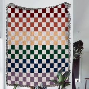 Könnte beinhalten: Eine gewebte karierte Wolldecke mit einem Regenbogenfarbverlauf. Die Decke ist an einer weißen Wand mit einem Fransenrand aufgehängt. Die Decke zeigt Farbtöne von Rot, Orange, Gelb, Grün, Blau und Lila.