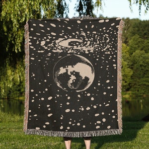 May include: A black woven tapestry with a large, silver disco ball in the center. The tapestry has a beige background with a swirling galaxy pattern and white dots.