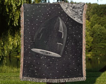 May include: A black woven throw blanket with a vintage illustration of a rocket ship traveling through space. The blanket features a white moon and stars on a black background. The text "the cool ruler co." is printed in white at the bottom of the blanket.