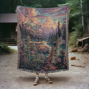 May include: A tapestry woven with a colorful landscape scene featuring a mountain range, a river, and a vintage yellow school bus. The tapestry is surrounded by green marijuana plants and has a fringe border.