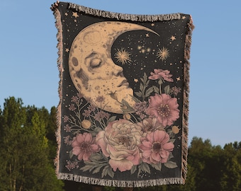 May include: A black woven tapestry with a crescent moon with a face and pink flowers. The moon is surrounded by stars. The tapestry is held up by a person in front of a blue sky and green trees. The text "the cool ruler.co" is at the bottom of the image.