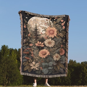 May include: A tapestry woven with a full moon, a forest scene, and a variety of flowers in shades of pink, yellow, and white. The tapestry is dark blue with a fringe border.