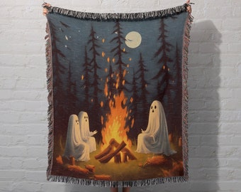 May include: A woven tapestry throw blanket featuring a whimsical scene of three ghosts gathered around a campfire in a dark forest. The moon is visible in the night sky, and the fire is glowing brightly.