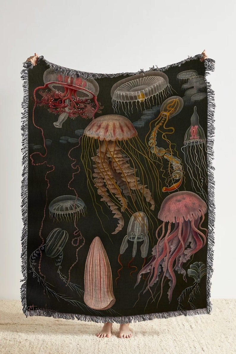 May include: A woven throw blanket with a dark background featuring a vintage illustration of various jellyfish in shades of pink, red, and yellow. The jellyfish are depicted with intricate details and are surrounded by a dark, swirling background.