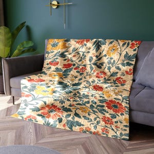 May include: A beige floral throw blanket draped over a gray sofa. The blanket features a repeating pattern of red, yellow, and teal flowers and leaves. The sofa is in front of a teal wall.