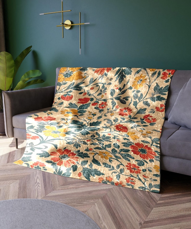 May include: A beige floral throw blanket draped over a gray sofa. The blanket features a repeating pattern of red, yellow, and teal flowers and leaves. The sofa is in front of a teal wall.